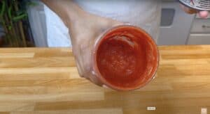 Person holding a blender cup with homemade tomato paste on a wooden countertop.